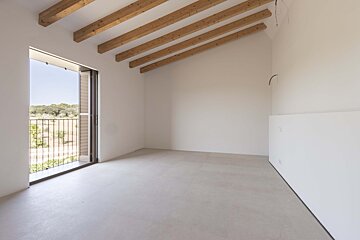Empty room with white walls, light floor, exposed wooden ceiling beams, and an open balcony door revealing a green, natural landscape.