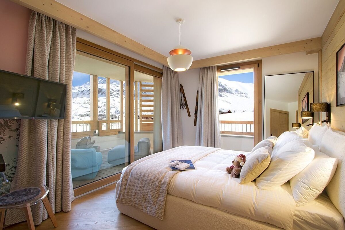 Modern ski chalet bedroom featuring a large bed, TV, and panoramic views of snow-covered mountains from expansive glass doors leading to a balcony.
