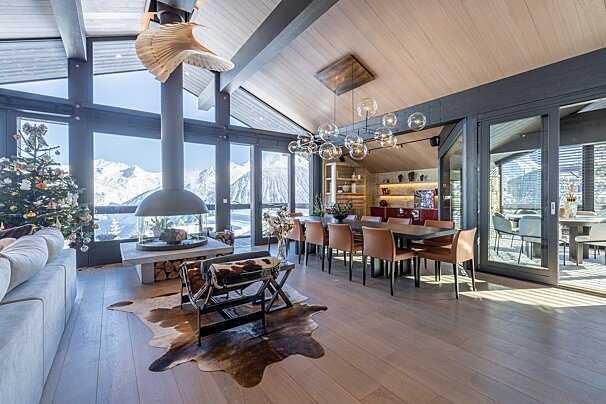 Luxurious chalet living/dining room with a Christmas tree, modern fireplace, and stunning snowy mountain views through large windows.