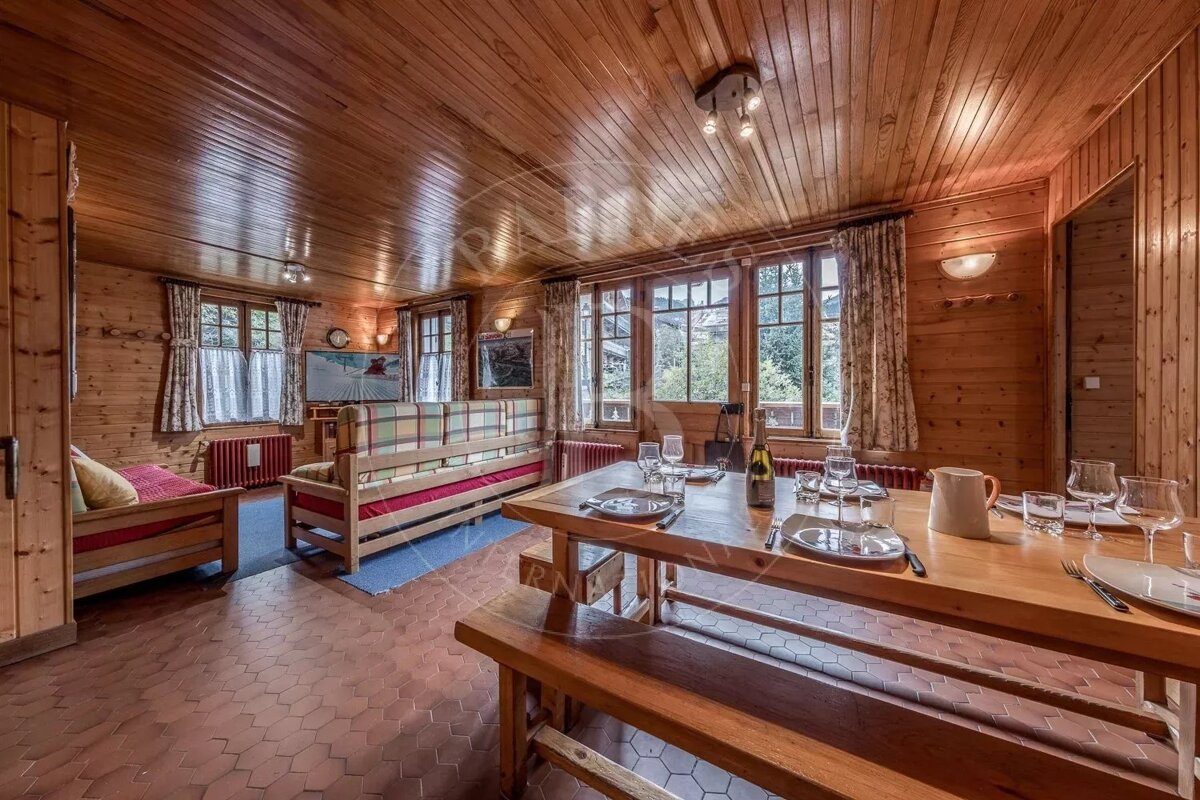 A rustic, wood-paneled room with a set dining table, benches, and cozy seating. Features large windows, warm lighting, and a hexagonal tiled floor.