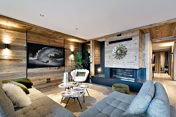A luxurious living room with abundant wood paneling, a modern fireplace, large TV, and comfortable seating, creating a warm, contemporary chalet ambiance.