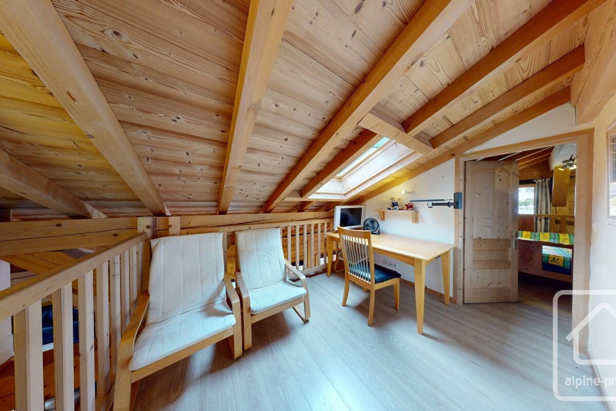 Rustic wood-paneled attic space with exposed beams, a skylight, desk, two armchairs, and a railing overlooking another room.