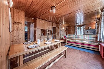 A warm, wood-paneled chalet interior with a dining table set for a meal, benches, a plaid sofa, and a terracotta hexagonal tiled floor.
