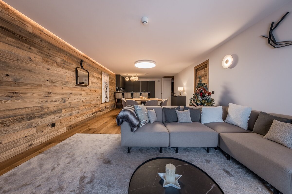 Cozy modern living room with a wood-paneled wall, large gray sectional sofa, Christmas tree, and an open dining/kitchen area.