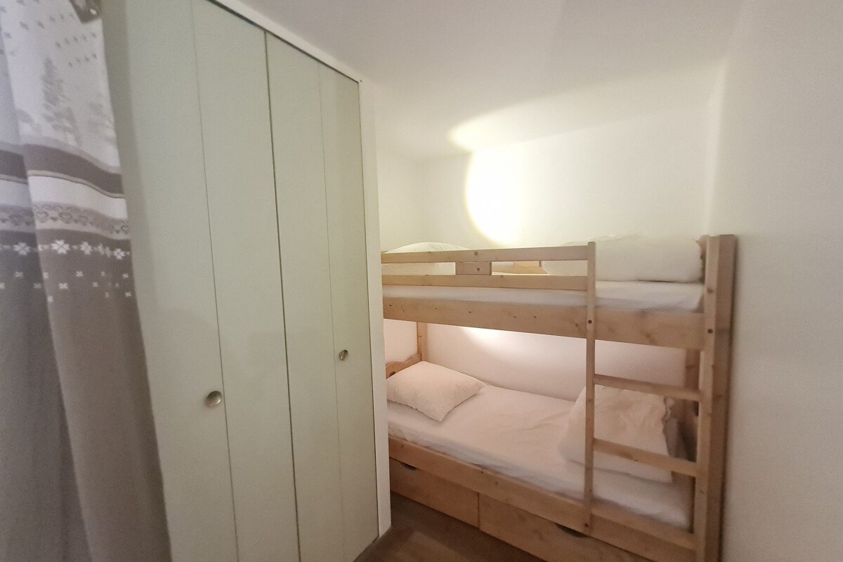 A cozy, compact room with light wooden bunk beds and white bedding. A light-colored closet and a patterned curtain are visible on the left.