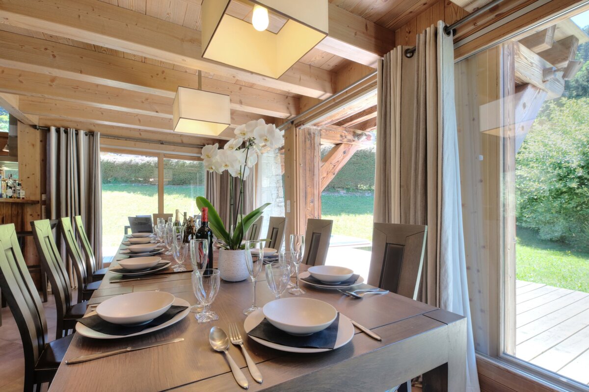 A rustic-chic wooden dining room with a long table set for a meal, featuring orchids, modern lights, and large windows opening to a lush green outdoor area.