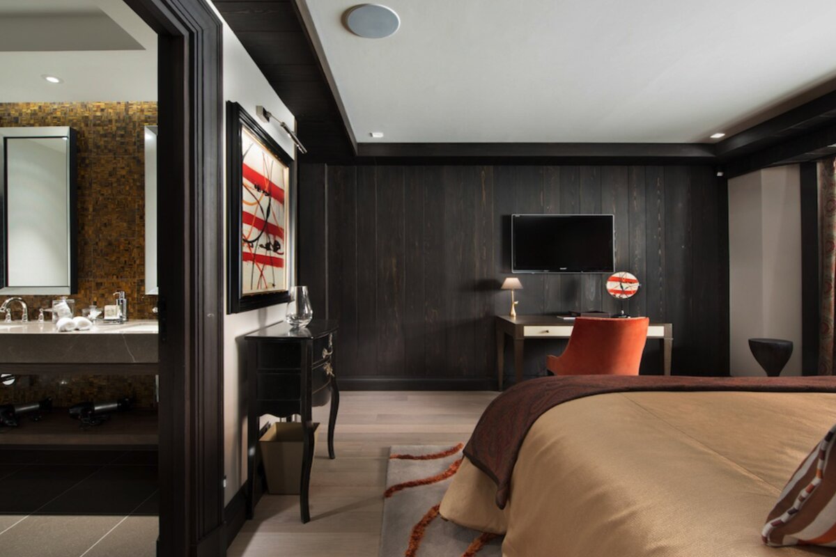 A modern bedroom featuring dark wood paneled walls, a desk with an orange chair, and an open view into an adjoining bathroom.