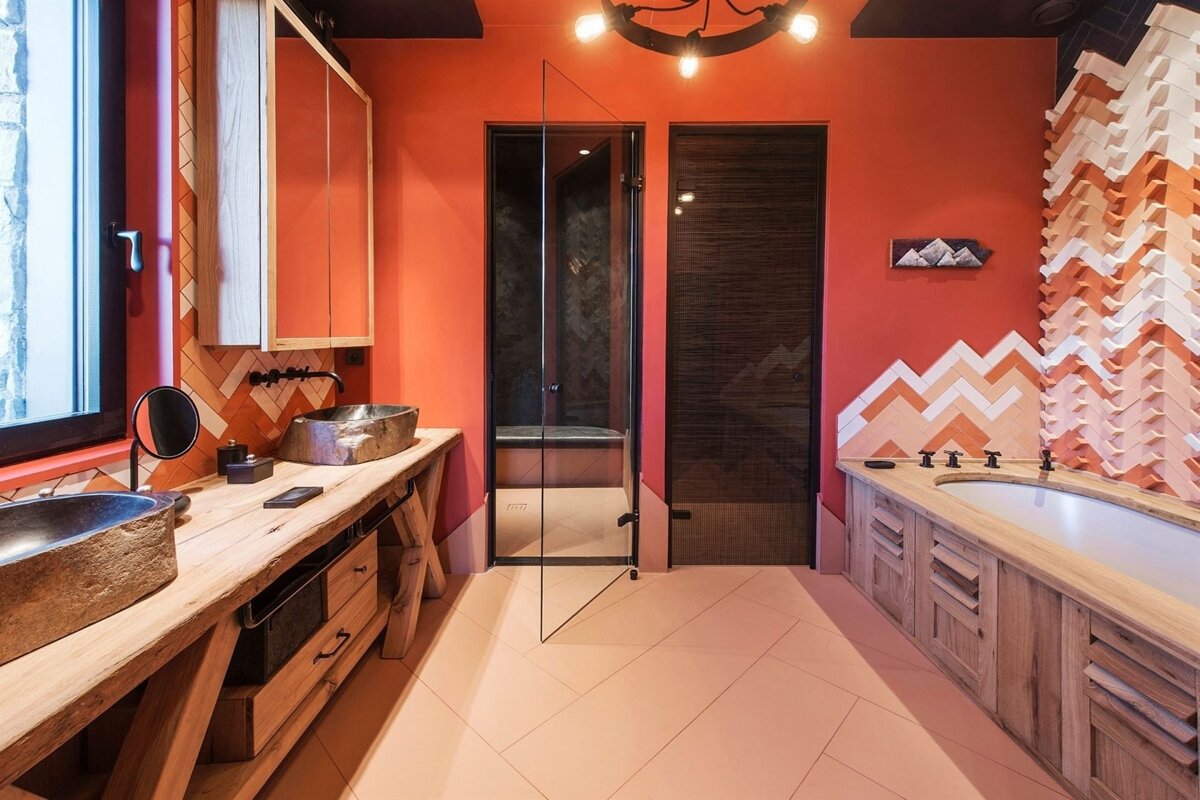 Vibrant orange bathroom with rustic wooden vanity, stone sinks, shower, and tub with striking chevron tile patterns, under industrial lights.