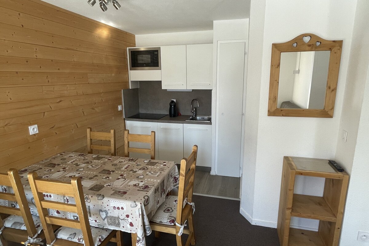 A cozy room with a dining table, wooden chairs, compact white kitchen with microwave, wood-paneled wall, and a heart-carved wooden mirror.