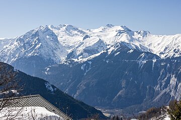 Snow-capped mountains tower over dark forested slopes under a clear blue sky, with a snowy roof visible in the foreground.