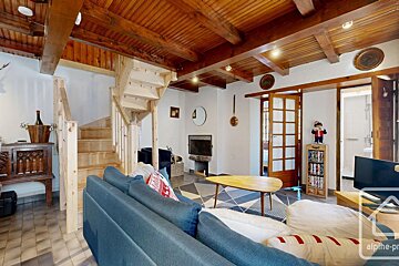 A cozy living room featuring a wooden beamed ceiling, light wooden staircase, blue sofa, fireplace, and patterned rug, with white walls.