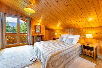 Inviting, all-wood bedroom with a cozy bed, warm lighting, and a charming balcony offering a scenic view of mountains and pine trees.