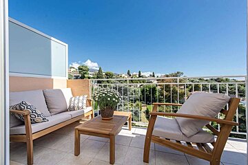 A sunny balcony with wooden outdoor furniture and a potted plant, overlooking a green landscape with distant buildings and mountains under a clear blue sky.