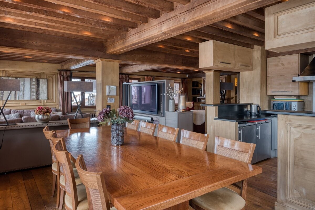 A cozy, open-plan wooden chalet interior with a long dining table, exposed beams, a living area with a sofa, and a modern kitchen visible.