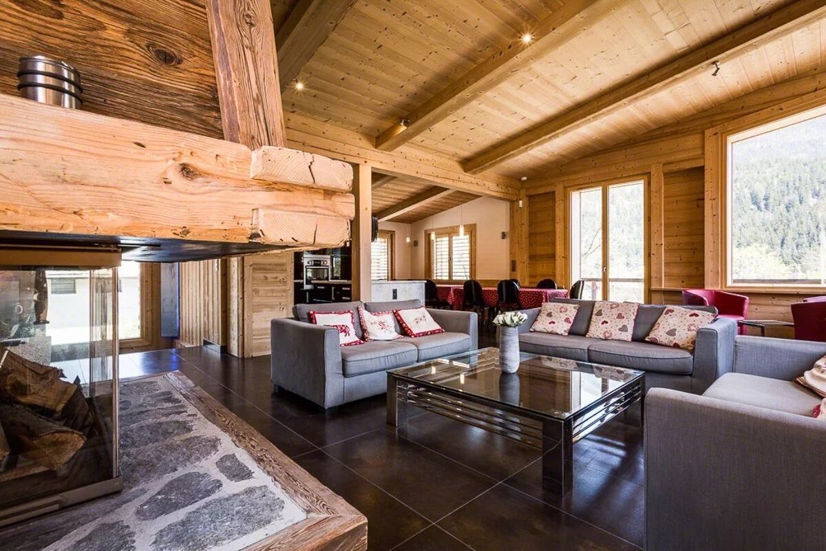 Cozy wooden chalet living room featuring grey sofas, a glass-top coffee table, and a large rustic fireplace.