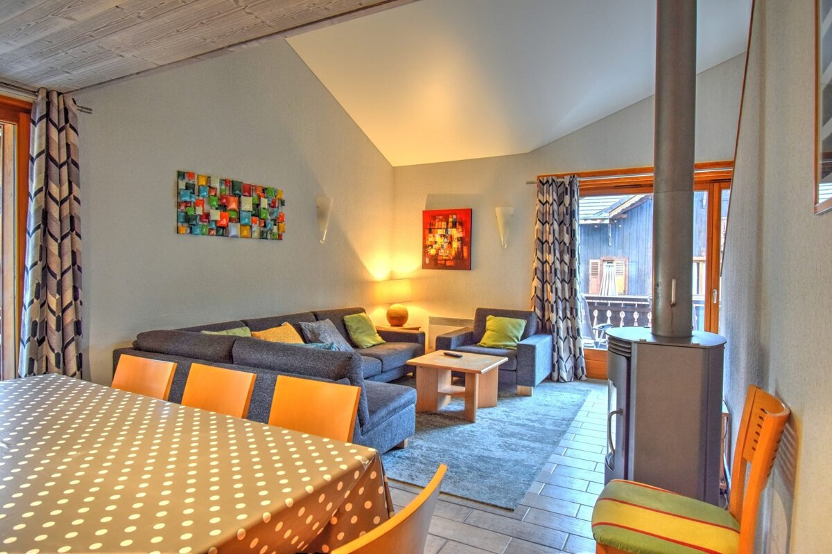 A modern, cozy living-dining room with a grey L-sofa, fireplace, dining table with polka dot cloth & orange chairs, and colorful art.