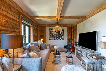 A rustic yet modern living room featuring warm wooden walls, ceiling beams, and a cozy gray sofa. A large TV, B&W art, and unique chandelier complete the inviting space.