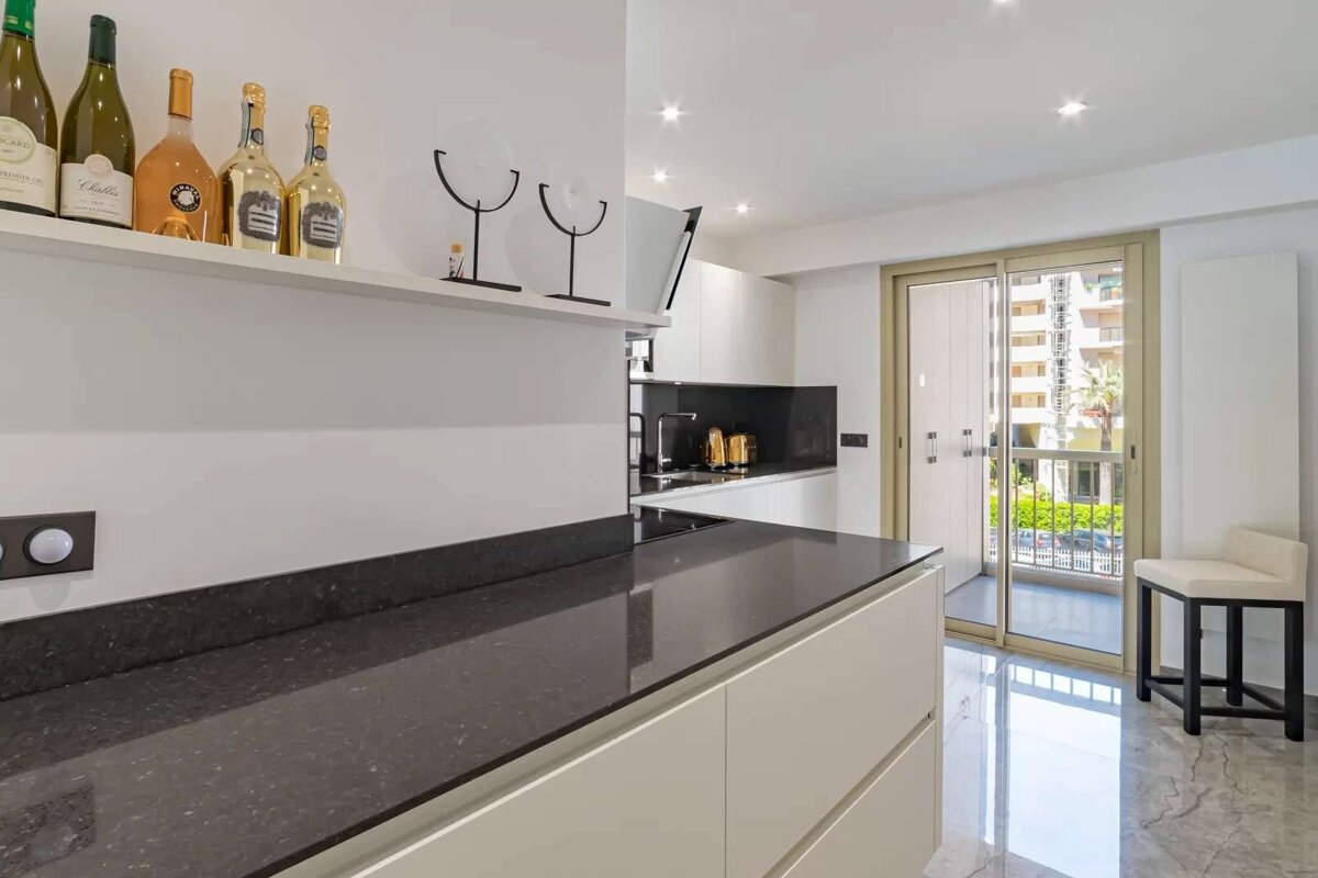 A modern kitchen features white cabinets, dark countertops, and bottles on a shelf. A sliding glass door opens to a balcony, and the floor is shiny.