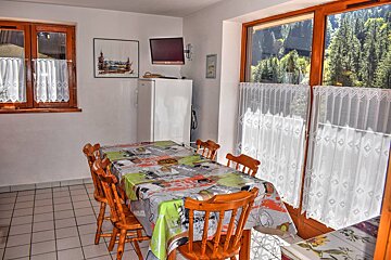 Cozy dining area with patterned table, wooden chairs, tiled floor, and windows overlooking a mountain forest. Fridge and TV are also present.