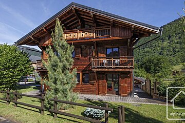A large wooden chalet with balconies, surrounded by green lawns, trees, and mountains under a blue sky.
