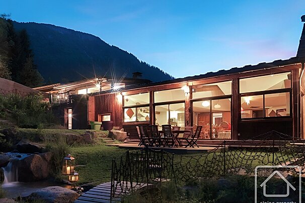 Dusk view of a mountain chalet with bright interior lights, an outdoor patio, a stream with glowing lanterns, and a dark mountain.
