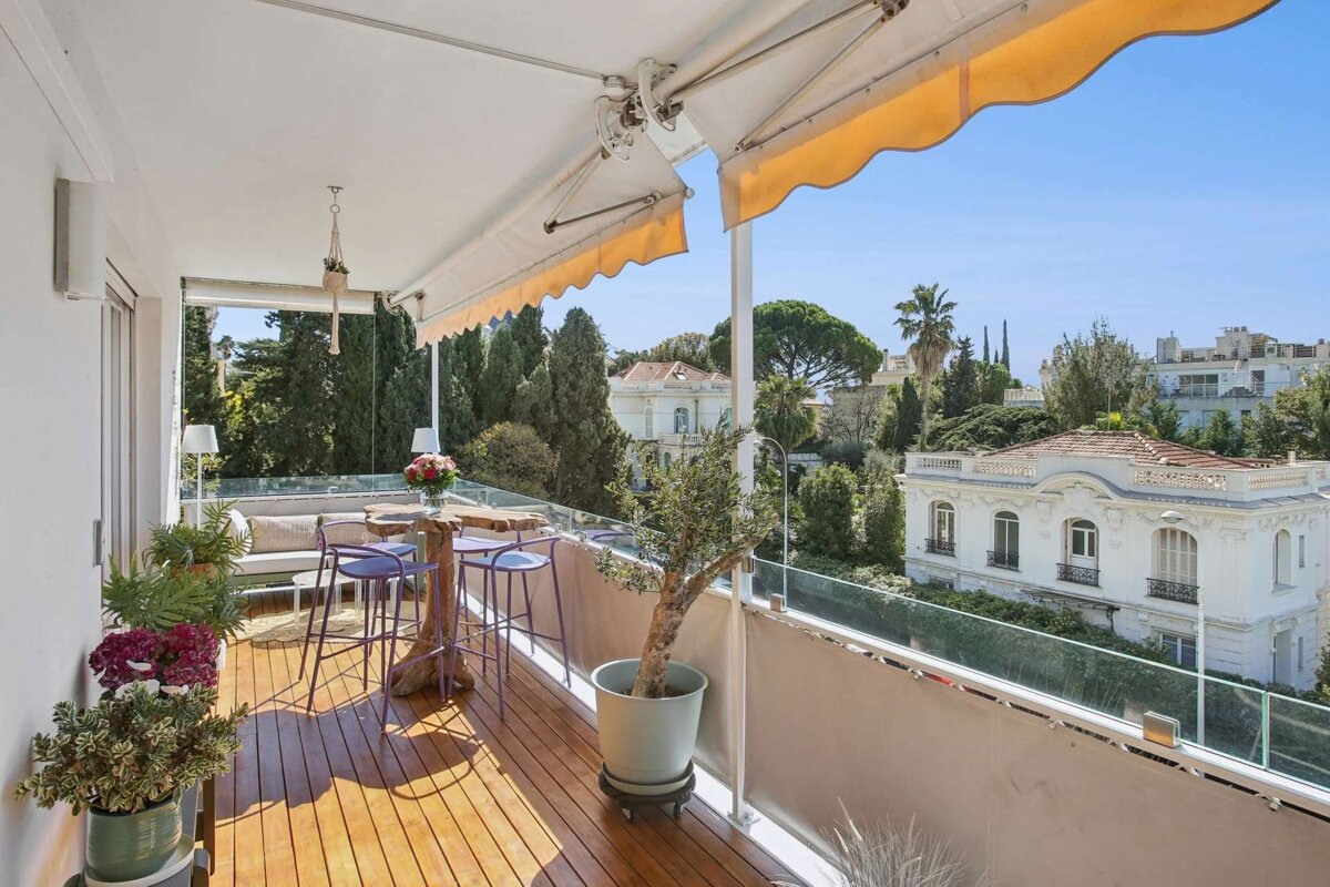 Sunny balcony with wooden floor, bar table, and plants, overlooking white houses and lush trees under a blue sky, shaded by an awning.