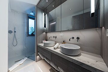 A luxurious modern bathroom featuring a double vanity with two vessel sinks, marble-patterned walls and counter, a glass walk-in shower, and ample lighting.