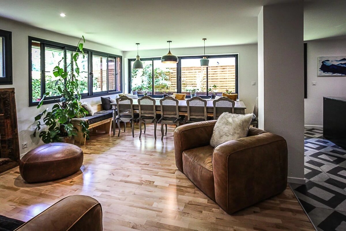 Spacious, bright living & dining room with warm wood floors, big windows, and comfy leather chairs. A large Monstera plant adds greenery.