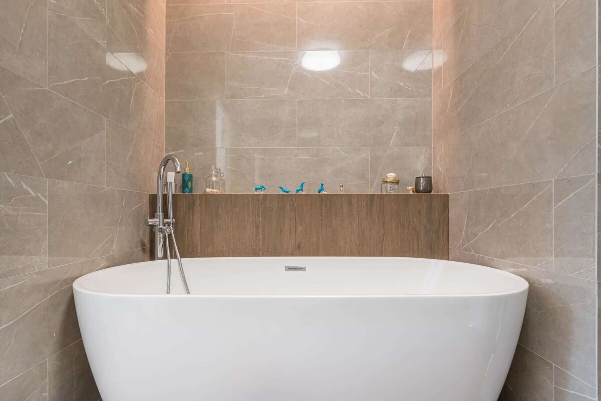 A modern bathroom features a white freestanding tub against grey marble-look tiled walls and a wooden shelf with decorative items.