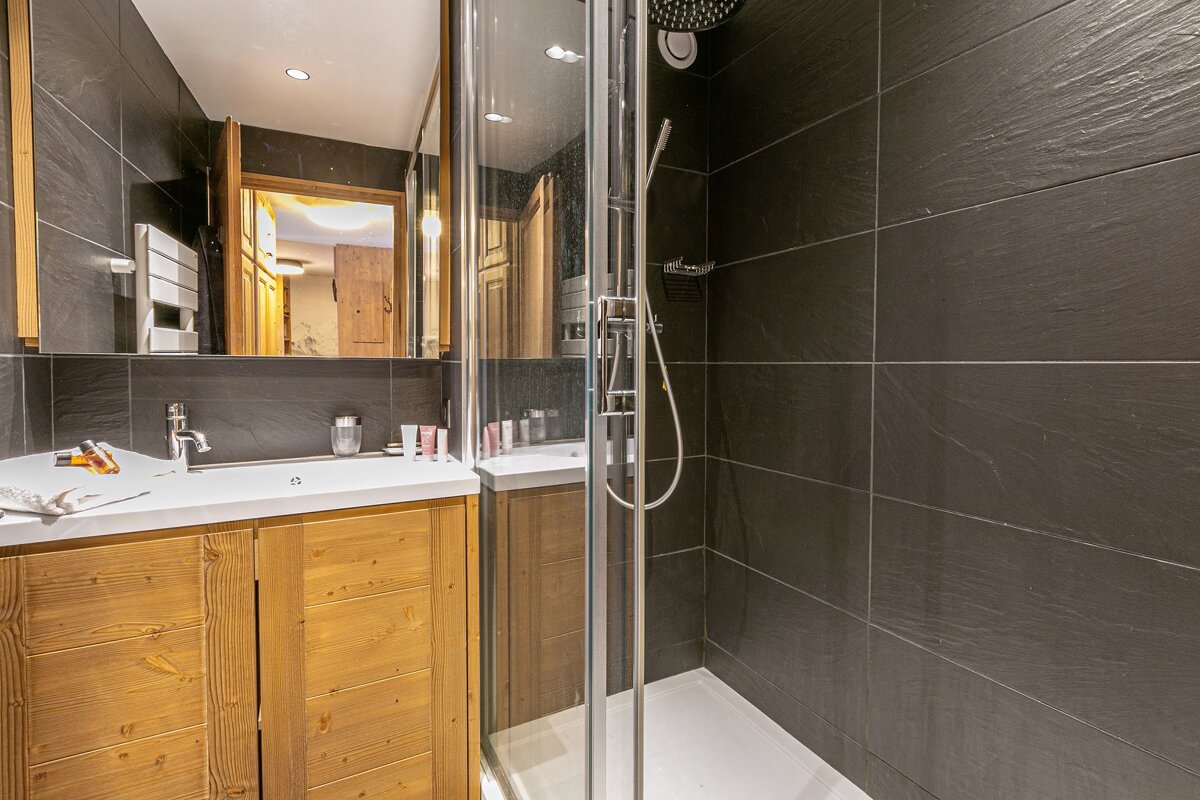 A modern bathroom with dark grey textured tiles, a wooden vanity with a white sink, and a glass shower enclosure featuring a rain shower head.