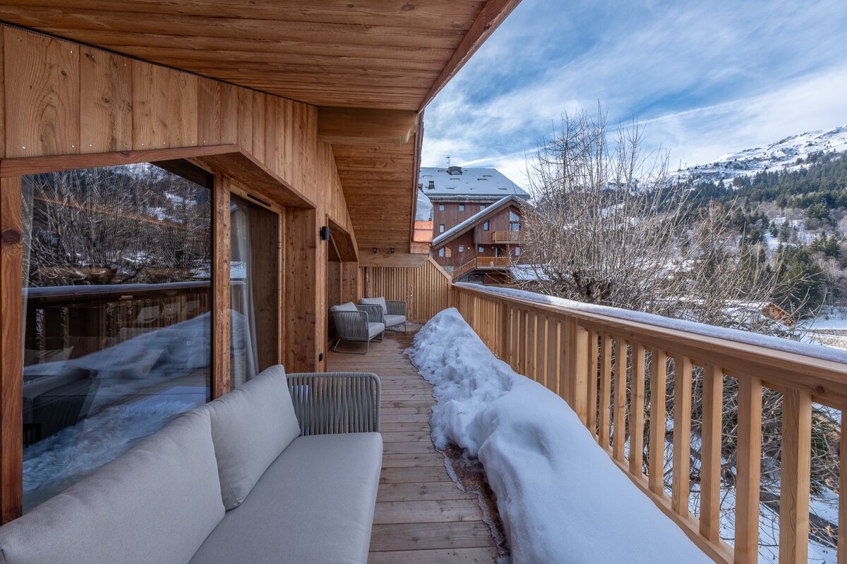 A cozy wooden balcony with outdoor furniture and a large snow pile, overlooking a snowy mountain landscape and other chalets under a blue sky.