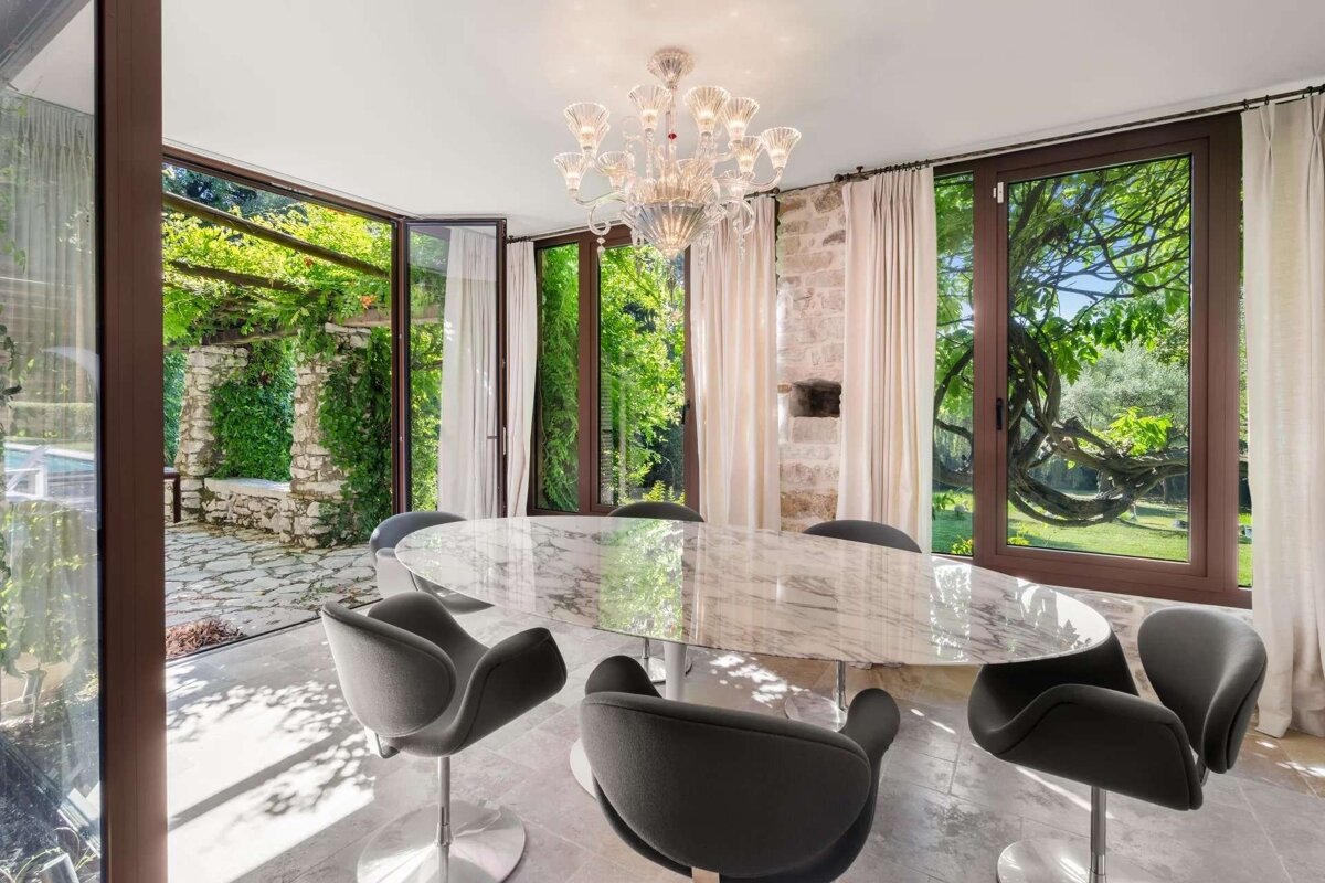 Bright, modern dining room with a marble table, stylish chairs, and a chandelier, offering expansive views of a lush green garden.