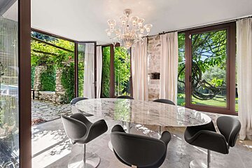 Bright, modern dining room with a marble table, stylish chairs, and a chandelier, offering expansive views of a lush green garden.