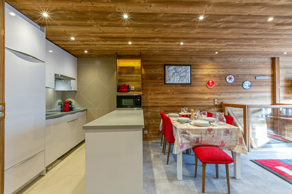 A modern, light-toned kitchen with white cabinets opens to a dining area featuring a set table with red chairs, all under a warm wooden ceiling.
