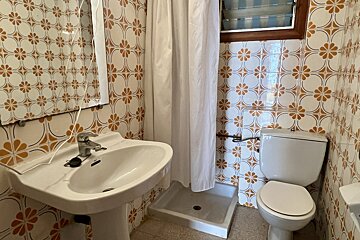 A small bathroom with brown and white patterned tiles, featuring a sink, toilet, shower with a white curtain, and a louvered window.