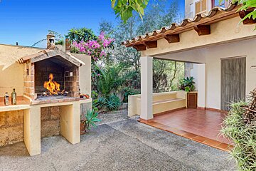 Sunny outdoor patio featuring a burning brick BBQ, a covered terrace with seating, and lush green garden foliage under a clear blue sky.