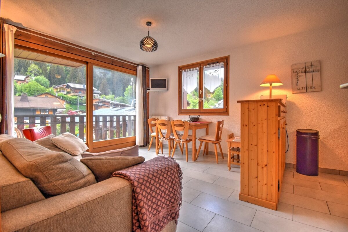 Cozy apartment interior featuring a comfortable sofa, wooden dining set, and large glass doors overlooking a scenic mountain village.