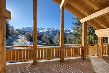 Sunny wooden balcony view of snow-capped mountains, a village road, and pine trees under a clear blue sky.