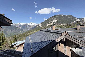Alpine village roofs overlook snow-capped mountains, green valleys, and a blue sky with white clouds on a sunny day.