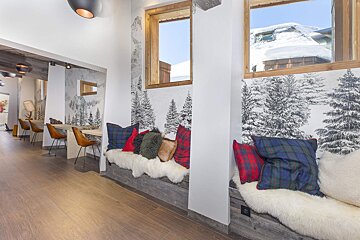 Cozy chalet interior featuring built-in window benches with plush sheepskins, colorful plaid pillows, and snowy forest wallpaper.