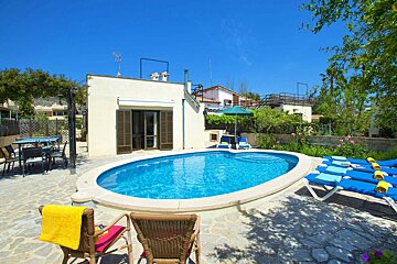 A sunny outdoor scene with a white house, an oval blue swimming pool, sun loungers, and an outdoor dining area under a bright blue sky.
