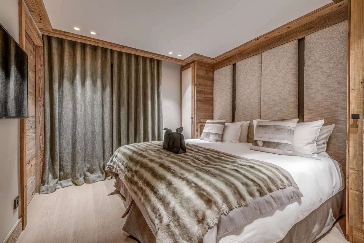 Modern, cozy bedroom with a large bed, faux fur blanket, wood paneling, grey curtains, and light flooring.