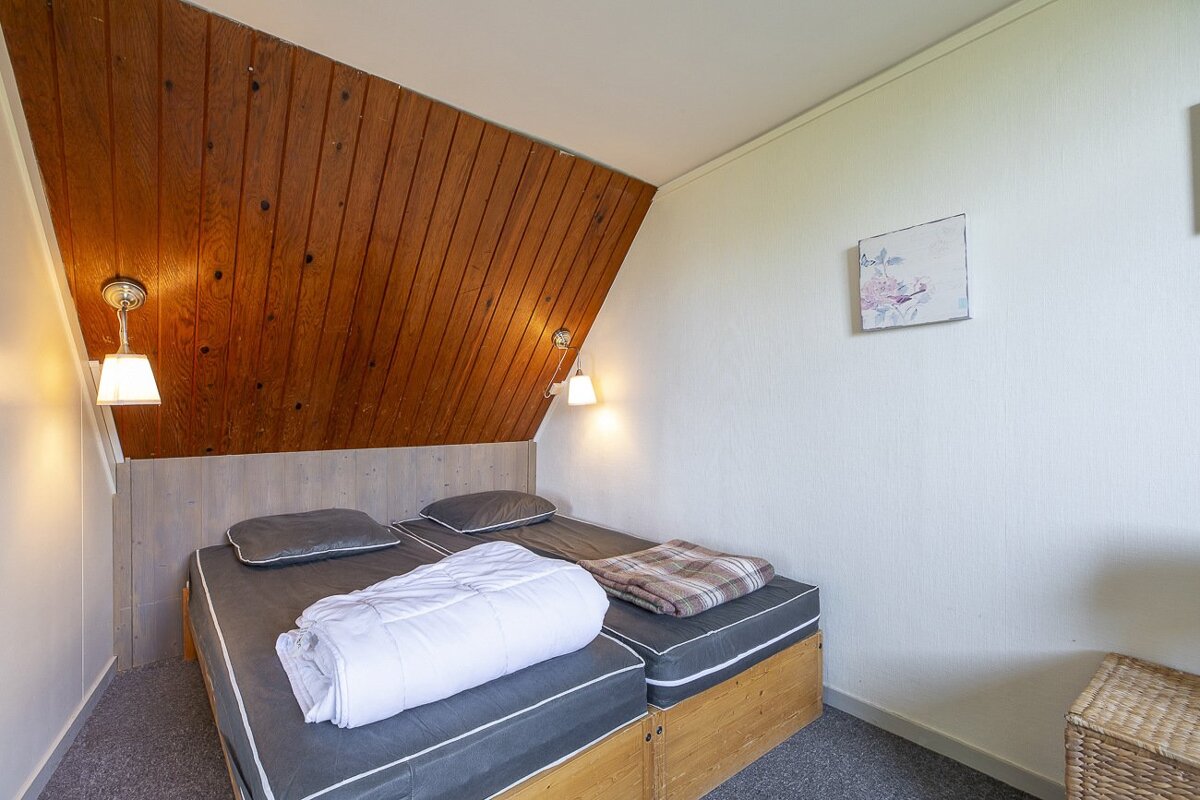 A cozy bedroom with two single beds, a sloped wooden ceiling, white walls, two wall lamps, and a small floral picture.