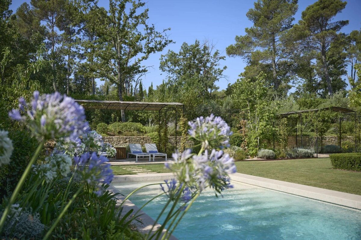 A tranquil garden scene featuring a blue swimming pool, lush greenery, a pergola, lounge chairs, and purple flowers in the foreground.