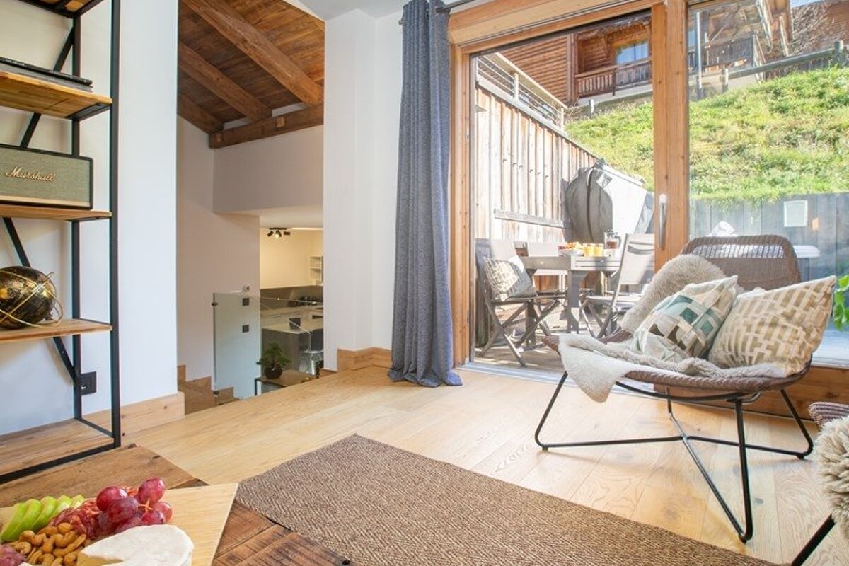 Bright chalet interior with light wood floors, a cozy lounge chair, and glass doors opening to an outdoor patio.