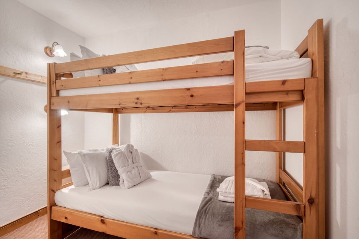 Wooden bunk beds with white linens, pillows, and folded towels in a simple white room.