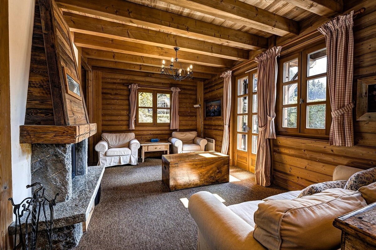 Warm, wood-paneled living room with exposed beams, stone fireplace, comfy sofas, and windows with plaid curtains, creating a cozy chalet ambiance.