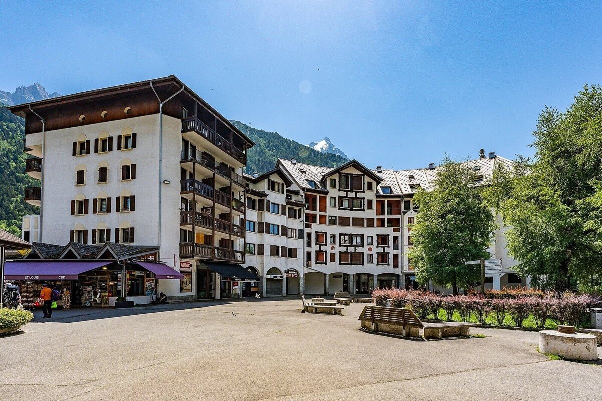 A sunlit alpine village square surrounded by chalet-style buildings, with forested mountains rising under a clear blue sky.