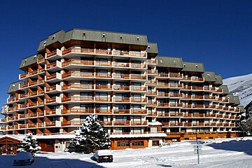 A large, multi-story terraced hotel with wooden balconies stands in a snowy landscape under a clear blue sky. Snow-covered trees and cars are visible.