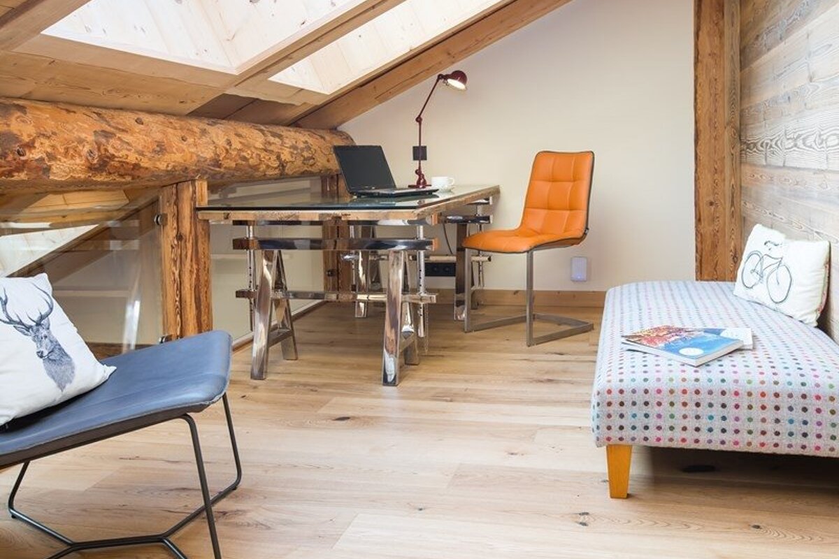Cozy attic workspace featuring a modern metal desk, orange chair, patterned daybed, and rustic wooden beams.